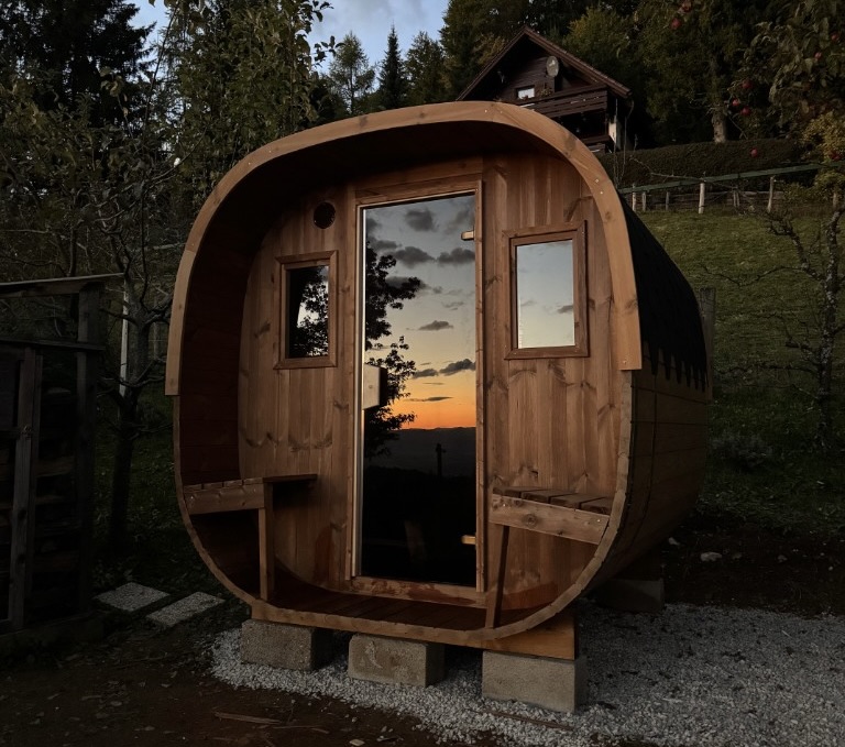 Zunanja sodna savna na betonskih blokih na prodnati podlagi, v ozadju hrib in gozd ob zahodu sonca.